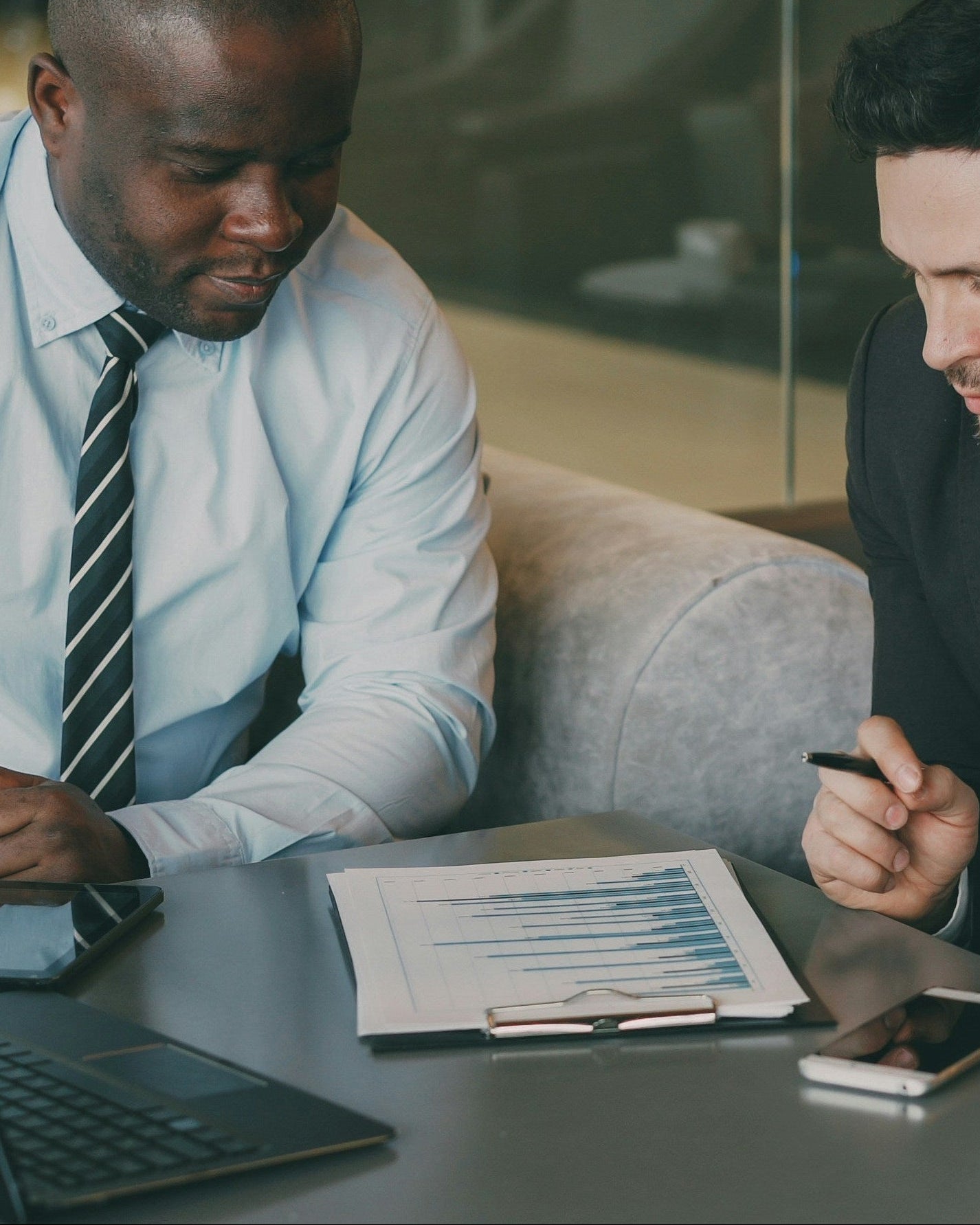 Two businessmen in a modern office setting, one using a laptop and the other holding a coffee cup.
